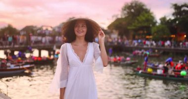 Beyaz elbiseli ve konik şapkalı beyaz kadın gün batımında Hoi An nehrinin kenarında gülümsüyor. Seyahat, yaşam tarzı, turizm tanıtımı veya romantik tatil reklamı için kavramsal görüntüler.