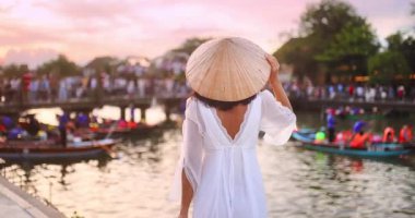 Geleneksel konik şapkalı beyaz elbiseli kadın gün batımında Hoi An nehrine bakıyor. Kamera yavaş hareket ediyor, manzaralı bir kültürel seyahat deneyiminin zarafetini ve sakinliğini yakalıyor..