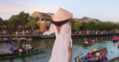 Beyaz elbiseli ve geleneksel şapkalı bir kadın Hoi An, Vietnam 'da nehirde tekne seyrediyor. Fenerler ve eski kasaba mimarisi ile huzurlu bir günbatımı sahnesi. Seyahat ve yaşam tarzı kullanımı.
