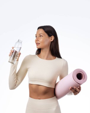 Elinde pembe yoga minderi ve su şişesi tutan bej renkli aktif giysili esmer kadın. Fitness, sağlık, yoga sınıfı tanıtımı, hidrasyon farkındalığı ya da sağlıklı yaşam tarzı reklamları için ideal..