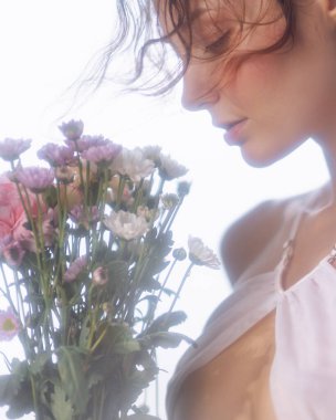 Beyaz iç çamaşırlı, yumuşak, romantik bir kadının portresi. Pastel çiçeklere bakıyor. Cilt bakımına, güzelliğe, moda editörlüğüne ya da duygusal hayat tarzına uygun..