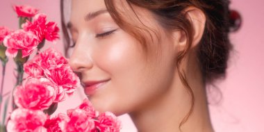 Kusursuz derisi ve kapalı gözleri olan, canlı pembe karanfillerin kokusundan zevk alan gülümseyen genç bir kadının yakın plan resmi. Pastel pembe arka plan ve yumuşak ışık rüya gibi bir ışık yaratır.