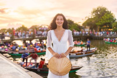 Uçan beyaz elbiseli gülümseyen Asyalı kadın güneş batarken Hoi An, Vietnam 'da nehrin kenarında elinde konik bir şapka tutuyor. Seyahat, yaşam tarzı, turizm, tanıtım ve kültürel konsept için ideal.