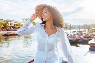 Vietnam 'da seyahat, kültür, moda ve yaşam tarzı tanıtım fotoğrafçılığı için ideal olan Hoi An' da fener teknelerinin yanında gülümseyen Vietnamlı koni şapkalı genç bir kadın..