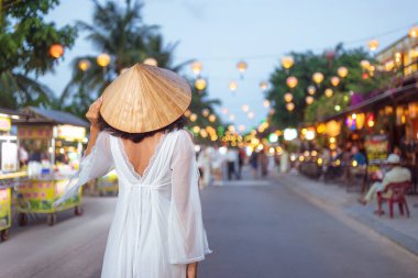 Beyaz elbiseli ve konik şapkalı bir kadının Hoi An, Vietnam 'da parıldayan bir fener sokağında yürüdüğünü gördüm. Seyahat yaşam tarzı tanıtımları, kültürel görseller veya moda yazıları için ideal.