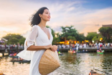 Güneş batımında seyahat reklamları, yaşam tarzı blogları ya da kültürel promosyonlar için ideal olan Hoi An, Vietnam 'da nehir kenarında elinde konik şapka tutan zarif beyaz elbiseli güzel bir Asyalı kadın..