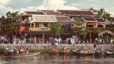 Hoi An, Vietnam 'ın manzara fotoğrafı, tarihi sarı binaların önündeki nehir boyunca renkli tekne ve kalabalıkları gösteriyor. Turizm, kültür makaleleri ve seyahat promosyonları için ideal.