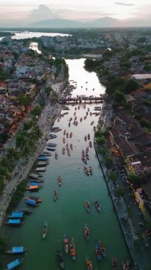 Pürüzsüz bir insansız hava aracı Hoi Nehri boyunca hareket eden bir görüntü yakalıyor. Altında tekneler, kıyıda turistler ve yukarıda günbatımı gökyüzü olan bir nehir. Seyahat, kültürel ve promosyon görselleri için harika..