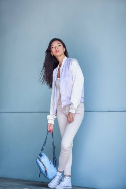 Pastel ceketli ve beyaz kotlu modaya uygun genç Asyalı kadın modern mavi bir duvarın önünde çantasıyla poz veriyor. Moda yazıları, yaşam tarzı markalaşması ve sokak tarzı kampanyaları için ideal..