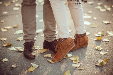 Romantik bir çift, sonbahar yapraklarıyla kaplı bir yolda birbirlerine yakın duruyor, rahat ayakkabılarını ve kotlarını sergiliyorlar, aşk, arkadaşlık ve mevsimsel açık hava aktiviteleri için mükemmel temaları var..