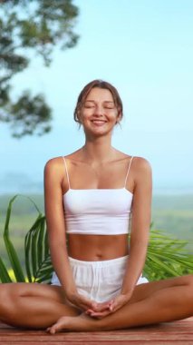 Doğayla çevrili, mutlu bir gülümsemeyle meditasyon yapan genç bir kadının dikey yavaş çekim videosu. Sağlık, farkındalık, yoga ve sağlıklı yaşam tarzı markalaşması için ideal..