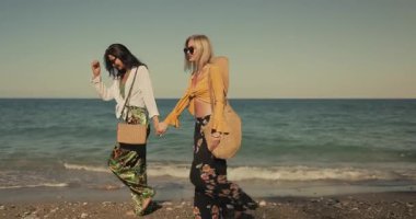 Renkli kıyafetler giymiş iki şık kadın güneşli bir plajda çıplak ayakla yürüyor, el ele tutuşuyor ve gülüyorlar. Arkadaşlık, yaz modası veya tanıtım içeriği için ideal yaşam tarzı ve seyahat görüntüleri.
