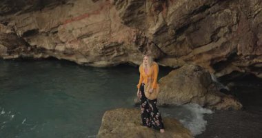 Çiçekli boho pantolonlu ve hardal bluzlu sarışın bir kadın deniz kenarındaki bir kayanın üzerinde dikiliyor ve uzaklığa bakıyor. Duygusal, moda, seyahat veya sağlık temalı sinematik görüntüler için ideal.