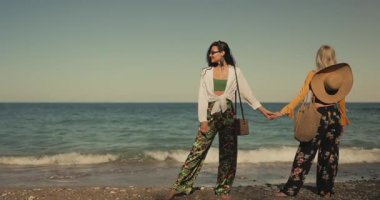 Boho kıyafetleri içinde iki şık arkadaş deniz kenarında keyifli bir anı paylaşıyor. El ele tutuşup kumsalda yalınayak sarılıyorlar. Yaşam tarzı, arkadaşlık, seyahat ve moda markalaşması için mükemmel..