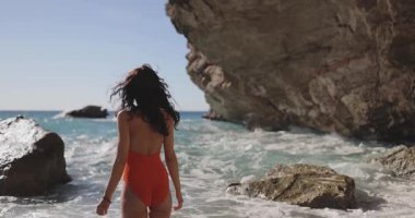 Kırmızı tek parça mayo giyen manken kıyı kayaları arasında denize doğru yürüyor, rüzgarda savrulan saçlar. Süper yavaş çekim görüntüleri lüks, güzellik, moda ve seyahat markaları için mükemmeldir..