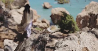 Şık bir kadın, turkuaz bir denize çıkan kayalık bir patika boyunca güneşli çam ağaçlarının arasında yürüyor. Seyahat, yaşam tarzı, macera ve doğa temalı ticari video içeriği için mükemmel.