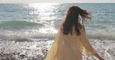 Yumuşak bej elbiseli genç bir kadın sakin bir plajda duruyor, sabah güneşi izlerken denize çakıl taşları atıyor. Düşünce, sağlık ve seyahat reklamları için mükemmel bir yavaş çekim klibi..