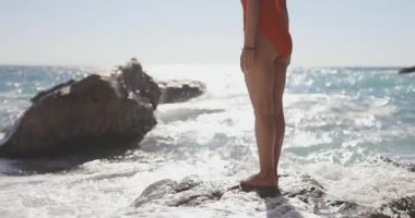 Şık bir genç kadın, etrafında dalgalar uçuşurken kırmızı mayo giymiş ıslak bir kayanın üzerinde cesurca poz veriyor. Süper yavaş çekimde çekildi, destansı, cesur ve güzellik kampanyaları için mükemmel..