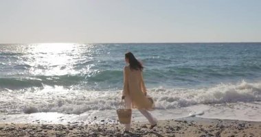 Modayı takip eden genç bir kadın kıyıda elinde kamış torbasıyla dalgaların altında sakince yürüyor. Yaşam tarzı, yaz modası ve sağlık temalı video kampanyaları için mükemmel..