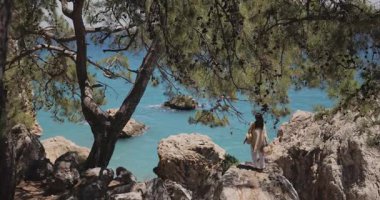 Huzurlu bir kadın, çam ağaçlarının çerçevelediği kayalık kayalıklarda sessizce duruyor ve canlı turkuaz okyanusa bakıyor. Çarpıcı geniş açı çekimi seyahat, doğa, yaşam tarzı veya refah içeriği için idealdir..