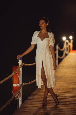 Beyaz elbiseli şık bir kadın gece Kemer, Türkiye 'de bir rıhtımda poz veriyor. Bu kaliteli görüntü, plajın zarafetini, güzelliğini ve dinginliğini yakalar. Moda, seyahat ve yaşam tarzı için mükemmeldir.