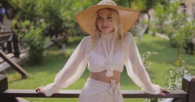 Şık yaz kıyafetleri ve geniş kenarlı şapkalı beyaz kadın güneşli bir bahçedeki ahşap korkuluklara yaslanıyor. Moda, yaşam tarzı, cilt bakımı veya tatil tanıtım içeriği için harika.