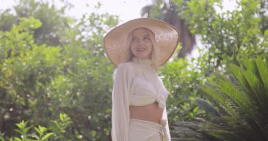 Son moda yaz kıyafetleri içinde şık beyaz kadın ve tropik bahçede açık havada poz veren geniş kenarlı şapka. Moda tanıtımı, tatil, yaşam tarzı veya güzellik reklamı kavramı.