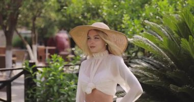 Beyaz düz bluzlu ve saman şapkalı güzel sarışın kadın yemyeşil bahçedeki ahşap terasta sakince poz veriyor. Yaşam tarzı, moda, güzellik veya daha etkili pazarlama için ideal.