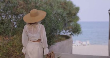 Beyaz takım elbiseli ve hasır şapkalı beyaz bir kadın zarif bir şekilde sakin bir plaja doğru yürüyor. Seyahat, tatil, moda ve zarif yaz atmosferli yaşam tarzı tanıtımı için ideal..
