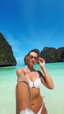 Tayland, Phi Adaları 'ndaki Maya Körfezi' nde güneşlenen beyaz bir kadın beyaz bir bikiniyle selfie çekiyor. Yaz modası, sahil seyahati ve etkileyici temalı videolar için mükemmel..