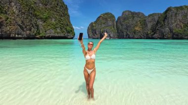 Tayland 'da Maya Körfezi' nde sığ turkuaz sularda selfie çeken bikinili bir kadın. Etkilendirici markalaşma, seyahat, yaz ve içerik oluşturucu yaşam tarzı video promosyonları için ideal.