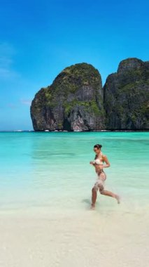Tayland, Maya Körfezi 'ndeki tropikal kıyı şeridi boyunca neşeyle koşan bikinili kadının dikey yavaş çekimi. Fitness, sağlık, seyahat ve yaz sahili yaşam tarzı video promosyonları için mükemmel..