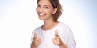 Kısa kıvırcık saçlı ve beyaz tişörtlü neşeli genç bir kadın, hafif eğimli bir arka plana karşı iki kez onay veriyor. Onun olumlu ifadesi ve onaylayıcı jesti güven ve...