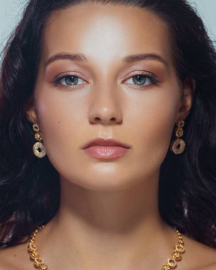 Kusursuz makyajlı, parlak tenli, zarif altın küpeli ve kolyeli bir kadının lüks stüdyo yakın çekimi. Lüks güzellik, moda ve cilt markası için ideal..