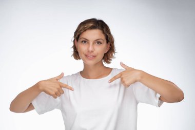 Kendine güvenen bir kadın boş beyaz tişörtüne işaret eder. Logo, mesaj ya da özel tasarım eklemek için mükemmeldir. Düz beyaz arkaplan ve net odak T-shirt 'ü promosyon için ideal hale getirir
