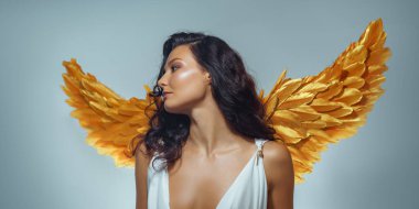 Altın melek kanatları ve derin kesimli beyaz elbisesi olan zarif bir kadın zarif bir bakışla poz veriyor. Moda, yayın ve tanıtım için ideal yüksek kalite fantezi güzellik görüntüsü.