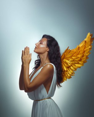 Beyaz elbiseli, altın kanatlı, gözleri kapalı ve elleri dua eden zarif bir kadının fantezi görüntüsü. Yumuşak ışık altında huzur içinde duruyor. Güzellik, manevi ya da sağlıklı bir promosyon için ideal..