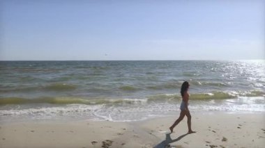 Güzel genç bir kadın, açık mavi gökyüzünün altında sahilde yalın ayak yürüyor. Seyahat, sağlık ya da yaz yaşam tarzı stok videoları ve yaratıcı tanıtımlar için mükemmel..