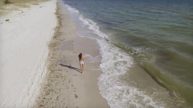 Sahilde yalınayak yürüyen genç bir kadının hava görüntüleri. Parlak güneş ışığı altında dalgalar geliyor. Seyahat, rahatlama, sağlık ve yaşam tarzı stok video kavramları için ideal.