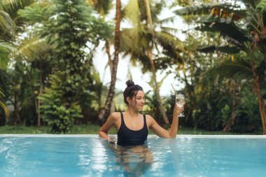 Siyah mayo giymiş kendine güvenen bir kadın, arka planda yemyeşil yemyeşil bir havuzda dinlenirken ferahlatıcı bir içecekten hoşlanır. Lüks, seyahat ve egzotik yaşam tarzı temaları için ideal.
