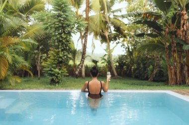 Mayo giymiş, elinde şarap bardağıyla havuzda gevşeyen tropikal bir ormana bakan bir kadının arka görüntüsü. Seyahat, tatil köyü, sağlık, spa ya da lüks eğlence deneyimleri için mükemmel..