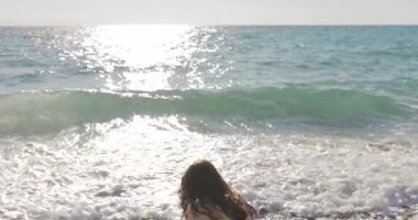 Okyanusa bakan kadın çakıl taşlarını nazikçe dalgalara fırlatıyor, güneşli bir sahilde huzurlu bir anın tadını çıkarıyor. Seyahat, farkındalık, doğa ve barışçıl yaşam tarzı içeriği için ideal.