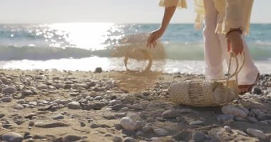 Kadın çantasını ve şapkasını güneşli bir çakıl taşı plajına koyar ve yumuşak sabah ışığında okyanusa doğru yürür. Kaçış temaları, farkındalık, boho yaşam tarzı veya barışçıl seyahat için ideal.