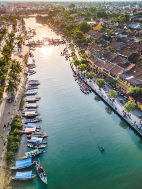Gün batımında Hoi An 'ın canlı nehir kenarı yaşamını, geleneksel tekneleri ve renkli tarihi caddeleri gösteren güzel drone görüntüsü. Turizm, seyahat yazıları ve tanıtım materyalleri için mükemmel.