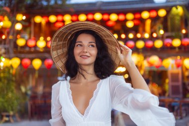 Konik şapkalı ve beyaz elbiseli kendine güvenen kadın Hoi An, Vietnam 'da parlayan fenerlerin altında gülümsüyor. Güzel bir yaşam tarzı ve seyahat fotoğrafı, başyazılar, reklamlar veya kültürel özellikler için mükemmel..
