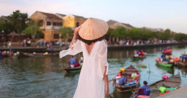Hoi An 'da nehrin kenarında duran zarif beyaz elbiseli ve konik şapkalı Asyalı kadının dikiz manzarası. Gün batımında kültür, kültür ve güzellikle huzurlu, şık bir seyahat tanıtımı..