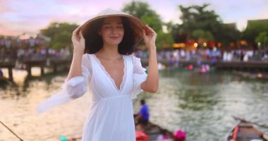 Beyaz elbiseli ve geleneksel şapkalı şık Asyalı kadın turist Hoi An, Vietnam 'da tekne gezisinde gülümsüyor. Kültürel reklamlar, moda veya lüks reklamlar için moda seyahat görüntüleri.