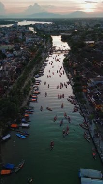 Hoi An Nehri 'nin günbatımında, geleneksel teknelerle ve nehir kıyısında çekilmiş dikey görüntüleri. Seyahat tanıtımları, Asya turizm içeriği ve atmosferik kültürel kavramlar için ideal.