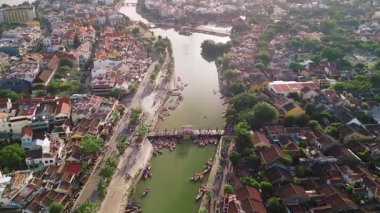 Hoi An 'ın manzaralı şehir manzarasından nehrin üzerindeki kalabalık yaya köprüsünün daha yakın görüntüsüne geçiş yapan yüksek kalite yatay drone görüntüleri. Seyahat ve turizm içeriği için ideal.