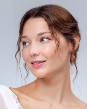 Gülümseyen genç bir kadının parlak yakın çekimi pürüzsüz, parlak bir teni ve yumuşak bir ifadesi var, yana bakıyor. Cilt bakımı, doğal güzellik, yaşam tarzı ve kozmetik reklamları için ideal..